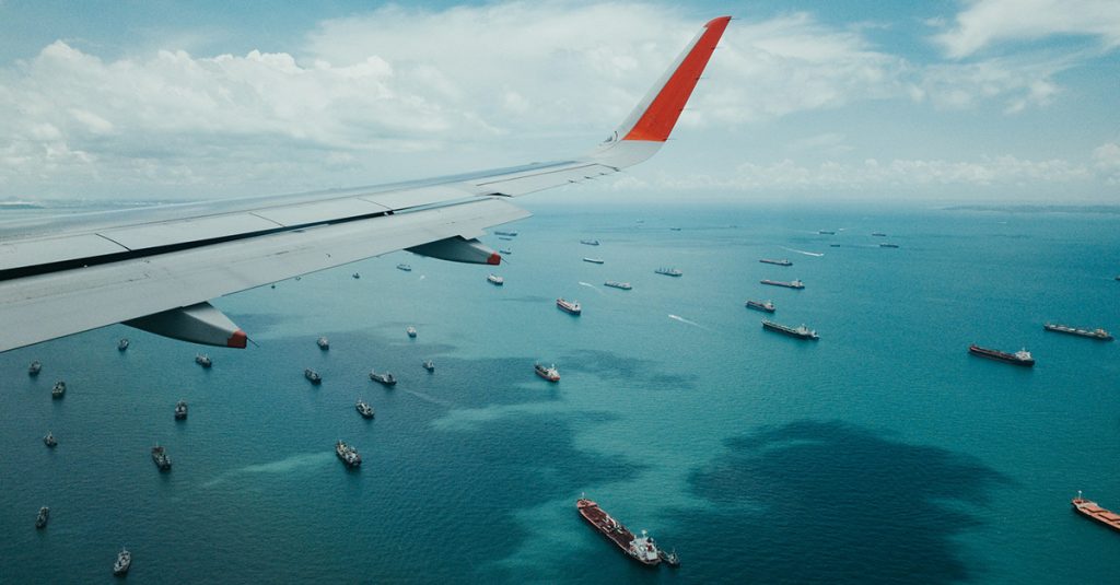 A photograph taken from the window of an aeroplane looking down on the ocean which features several container ships.