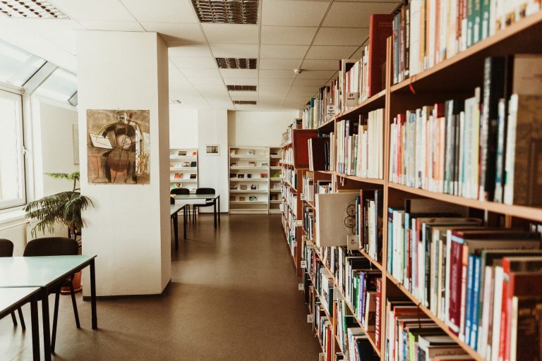 A photo depicting a university library with a bookshelf on the right hand side and some tables on the left.